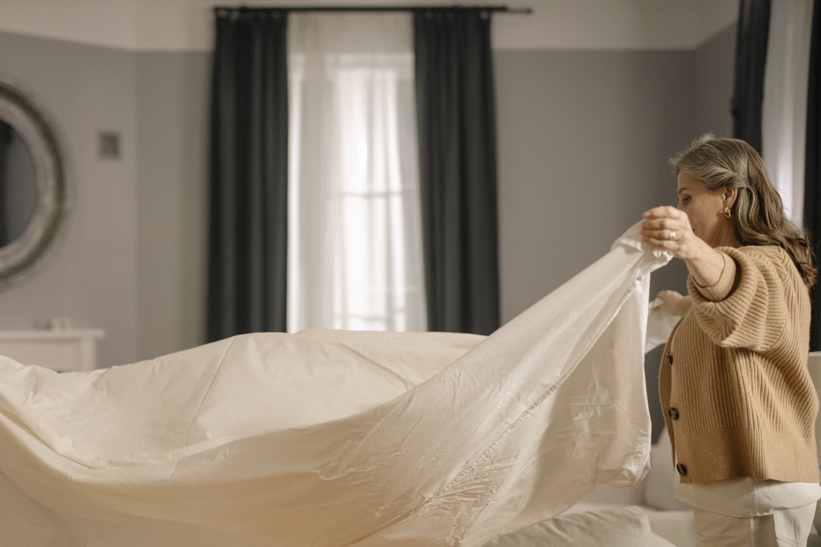 A person spreading a fresh, clean white sheet over a bed in a softly lit bedroom.